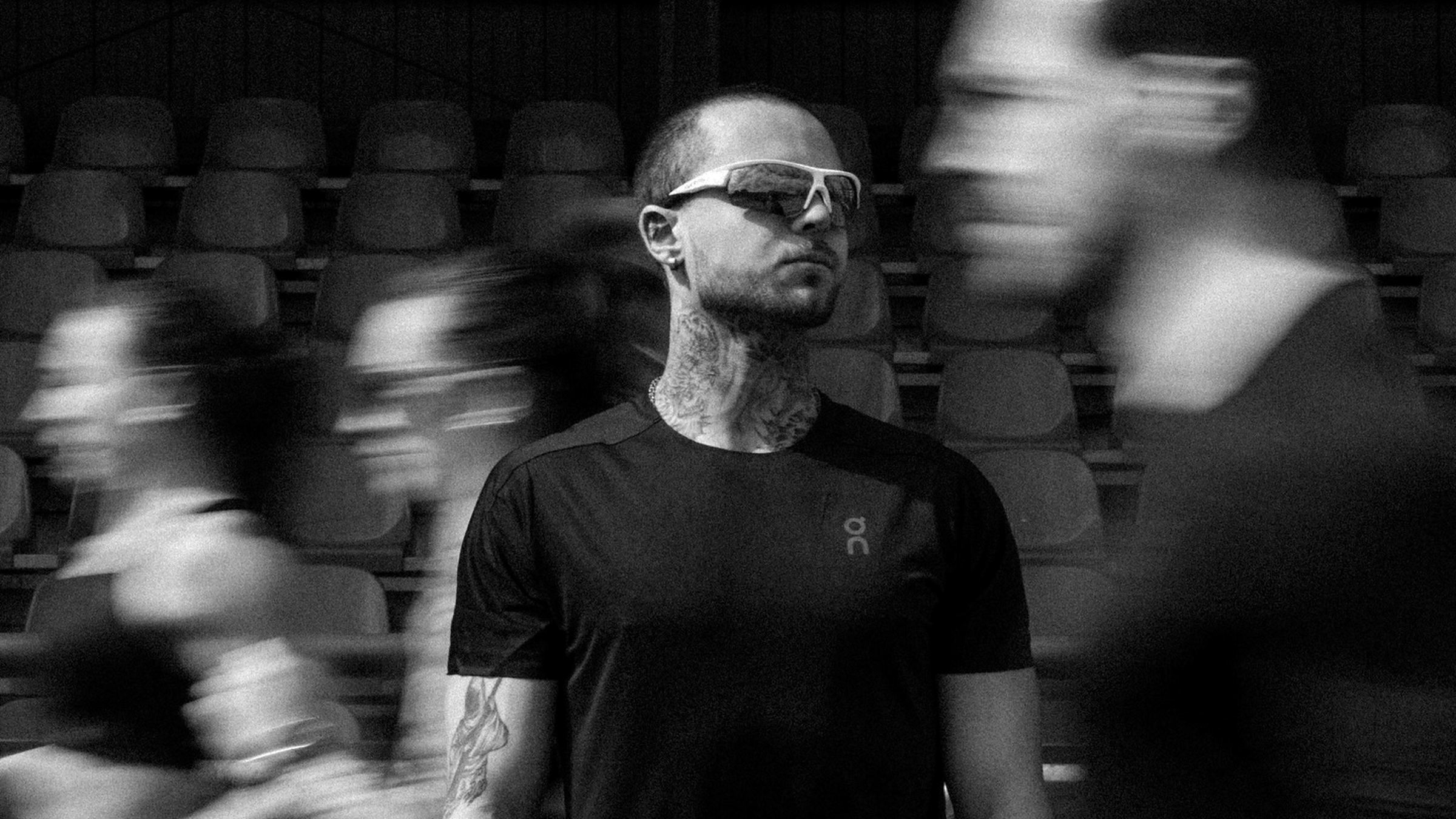 Oliwier Olszewski stoi nieruchomo na stadionie, obok przebiegają rozmyci biegacze, sesja zdjęciowa atletów nuvr, kwiecień 2026